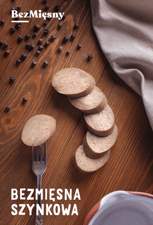 Packshot of Bezmięsna szynkowa: plant-based ham slices on a dark wooden table strewn with peppercorns, one slice held up on a fork