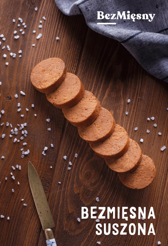 Packshot of Bezmięsna suszona: a row of plant-based dry sausage slices on dark wood scattered with coarse salt, a knife alongside
