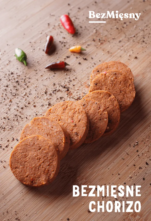 Packshot of Bezmięsne chorizo: plant-based chorizo slices fanned out on a light wooden board with chilli peppers and cracked pepper