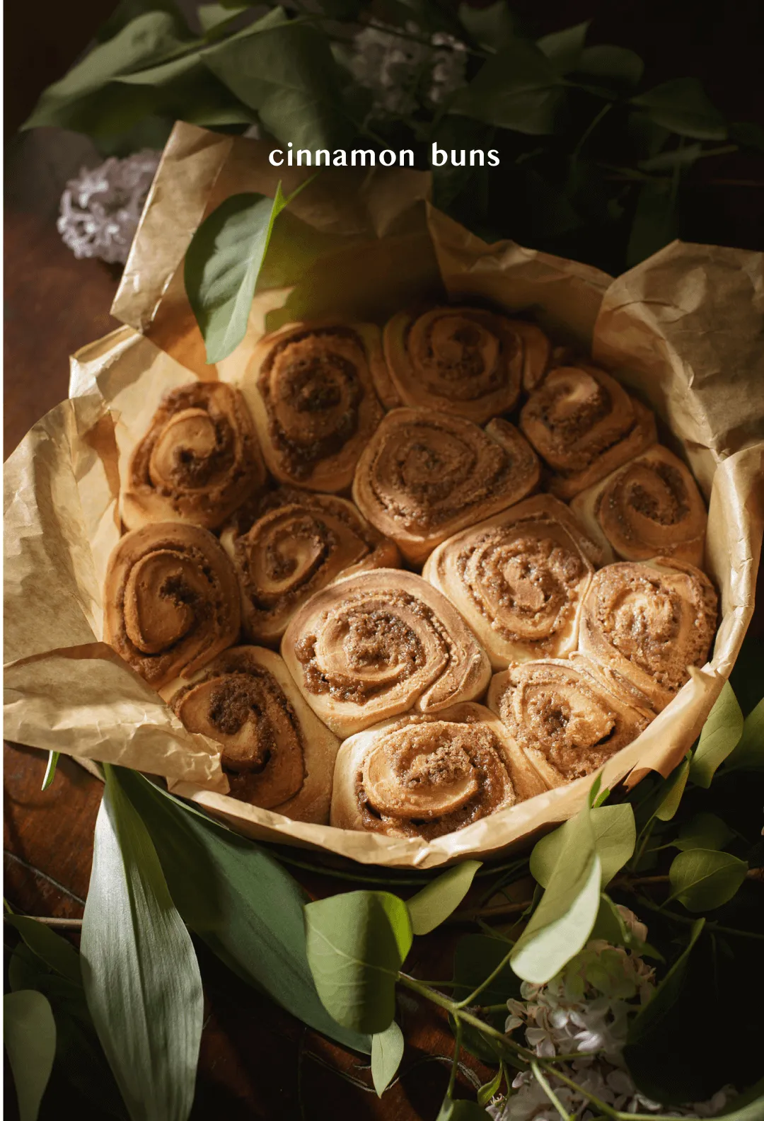 Pan of freshly baked cinnamon buns on baking paper surrounded by eucalyptus, titled "cinnamon buns"