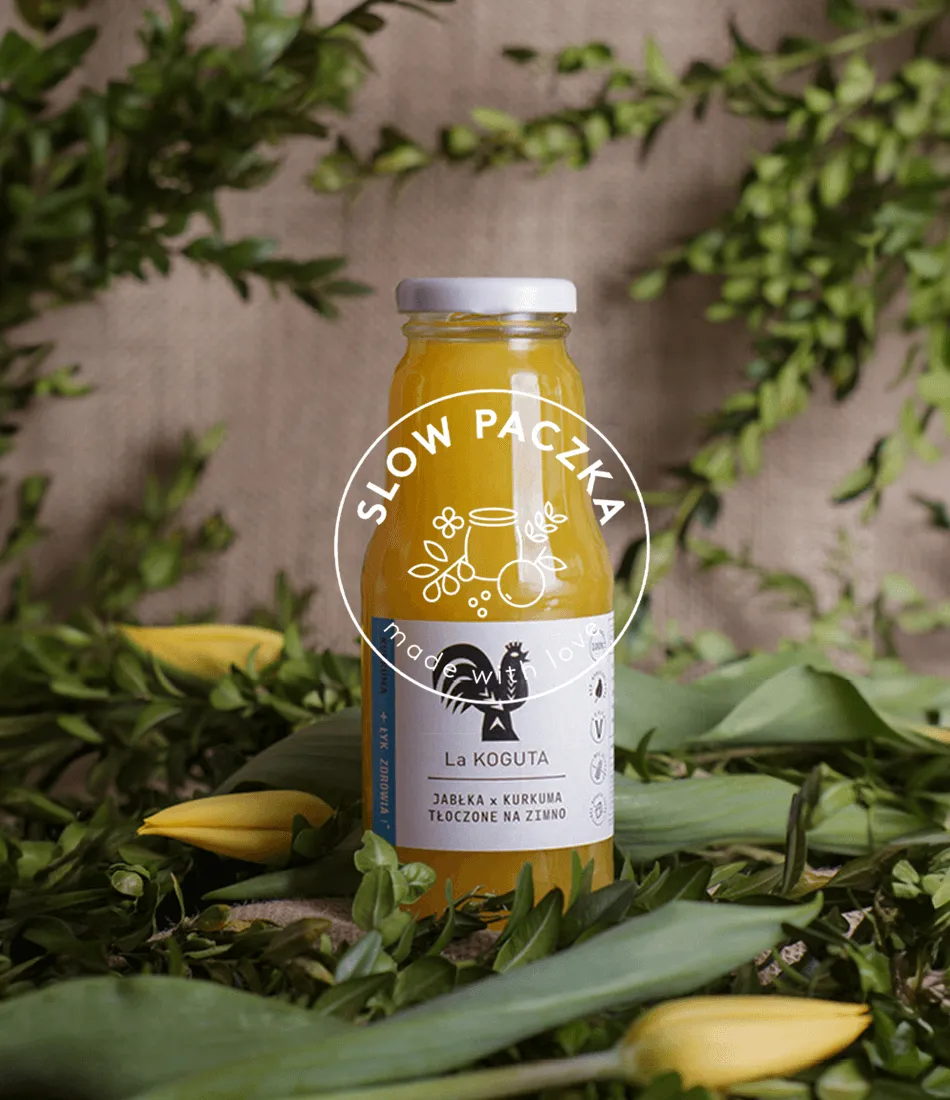 Bottle of La Koguta apple and turmeric juice among greenery and yellow tulips, with the round Slow Paczka "made with love" emblem overlaid