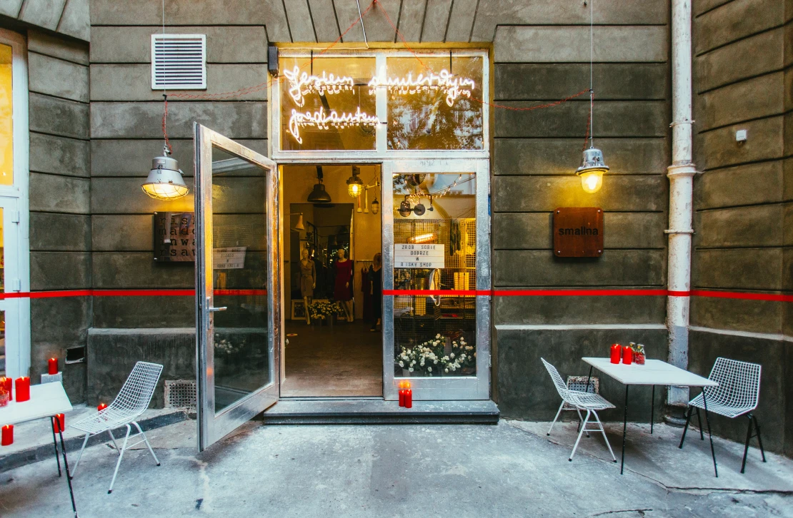 Courtyard entrance to the RISK boutique, with a neon sign in the window, a red tape line across the facade, and string lights and candles over outdoor café tables