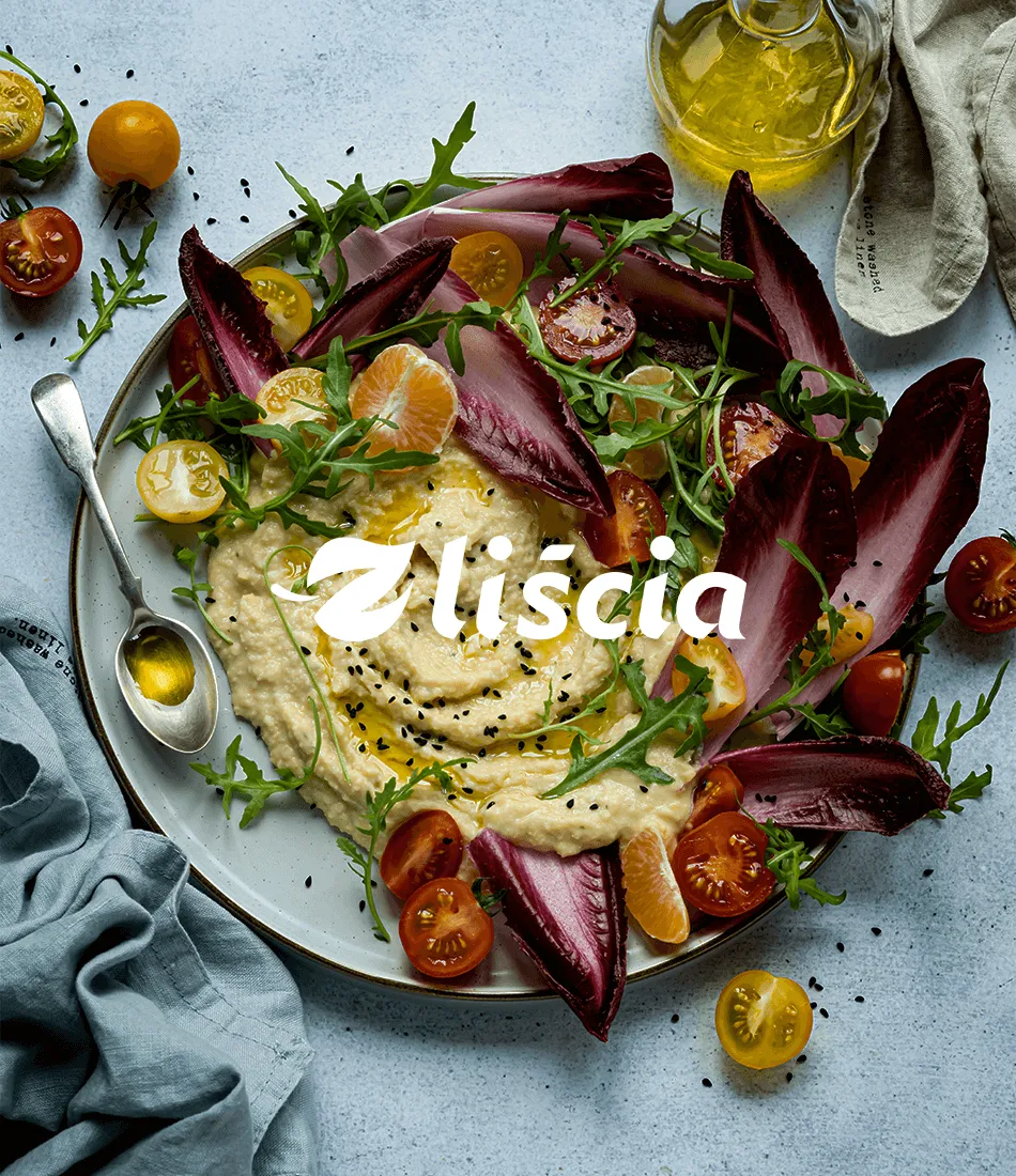Overhead photo of a plate of hummus topped with red chicory leaves, cherry tomatoes and rocket, with the Z Liścia logotype overlaid